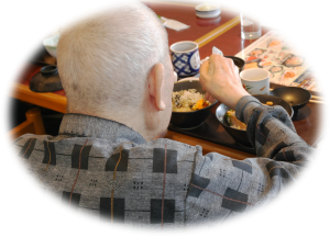 高齢者が食事をしている様子。テーブルで食器を使用している生活風景。